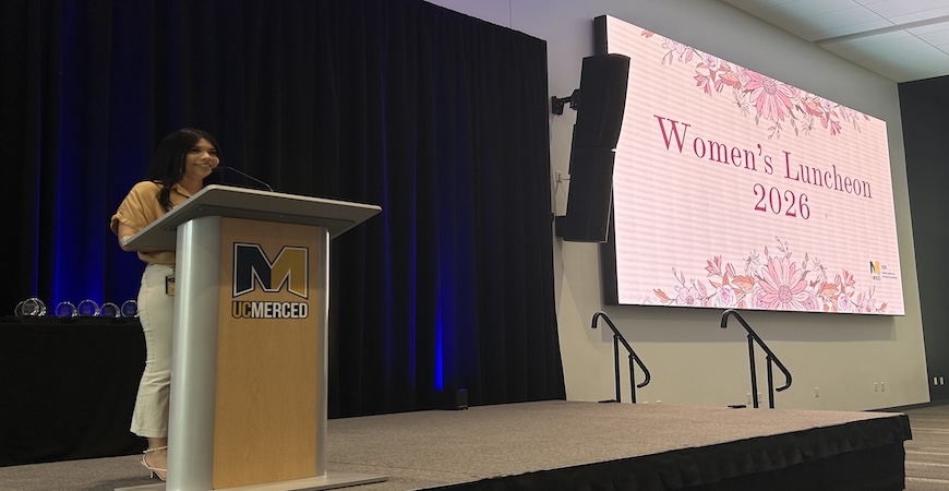 EJIE Student Success Manager Lorena Mejia stands at a podium. A pink slide that reads Women's Luncheon 2026 is projected to the right.