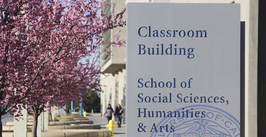 A photo of the Classroom Building that houses the UC Merced School of Social Sciences, Humanties, and Arts. Cherry blossoms bloom to the left of the sign.